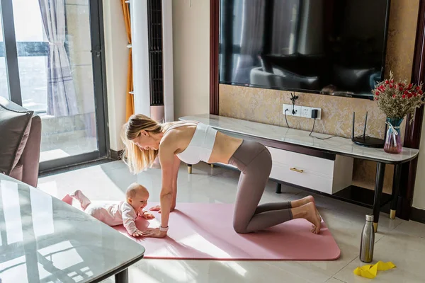 赤ちゃんと一緒に家で運動するスポーツウェアの若い母親。コロナウイルスcovid-19隔離中のオンライントレーニング。パンデミック・ロックダウンの間も安全と安定を保ちなさい。スポーツ、フィットネス、健康的なコンセプト ロイヤリティフリーのストック画像