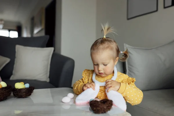 Niña caucásica feliz de un año en casa en la sala de estar con huevos de Pascua de colores. Permanecer en casa durante la pandemia del Coronavirus covid-19 — Foto de Stock