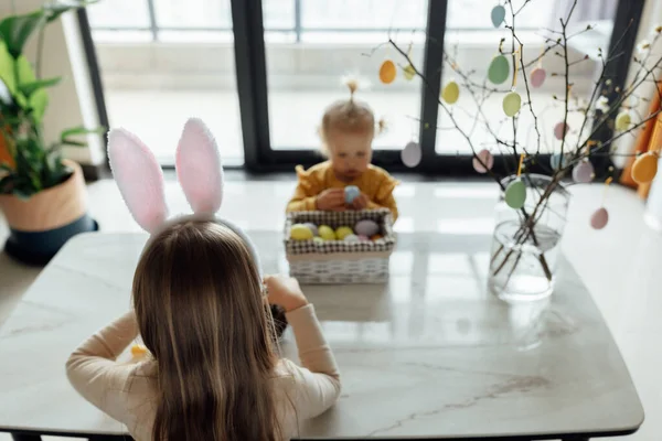 Niña caucásica feliz niño de ocho años y su hermana menor de un año en casa en la sala de estar con huevos de Pascua de colores. Permanecer en casa durante la pandemia del Coronavirus covid-19 — Foto de Stock