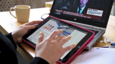 Siyah elbiseli bir kız elinde tablet tutarak ders çalışıyor, bir dizüstü bilgisayardan aşı haberlerini izliyor. Yakın plan.