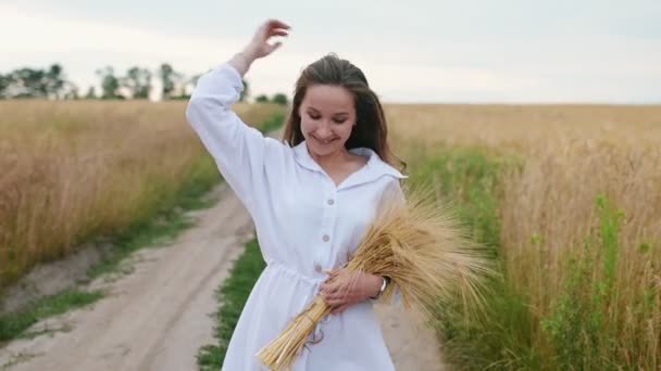 Genç, mutlu bir kadın buğday tarlasında yürüyor elinde buğdayla gülümsüyor, elinde buğday tutuyor, sinematik sabah çekimi, ağır çekim, 4k, yakın çekim. — Stok video