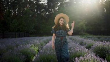 Elbise üzerindeki çekici kıvırcık kız, geniş kenarlı bir yaz şapkasıyla kameraya poz verirken mutlu bir gülümseme, lavanta sahasında yürüme, 360 kamera efekti, Girdap, Ağır çekim