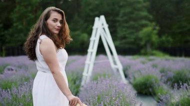 Güzel kıvırcık kadın lavanta tarlasında ya da sokakta yürüyor, elinde şapkasıyla kameraya poz veren açık renk bir elbise giyiyor ve güzel bir gülümseme sergiliyor, rüzgar saçlarıyla oynuyor. Açık havada.