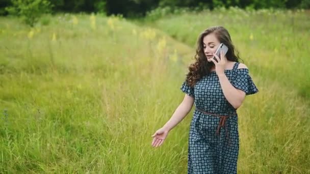 Mujer caucásica joven confiada y sonriente sosteniendo el teléfono durante el caminar hablando con el mejor amigo en el teléfono móvil en el tiempo soleado ventoso en el paisaje en la aldea, cámara lenta — Vídeo de stock