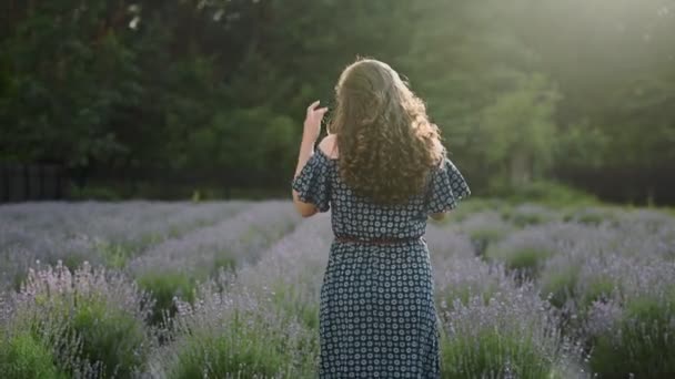 Lockiges brünettes Mädchen geht in einem Lavendelfeld, lächelt, blickt in die Kamera, Rückansicht, sonniges Wetter, Zeitlupe, Nahaufnahme — Stockvideo