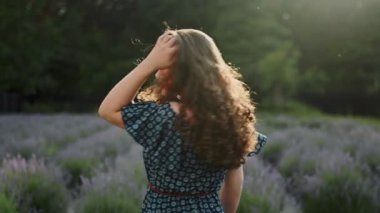 Çekici kıvırcık esmer kadın lavanta çalılarının arasında yürüyor, beyaz mutlu, kameraya bakıyor lavanta rengi arka manzara, güneşli hava, yavaş çekim