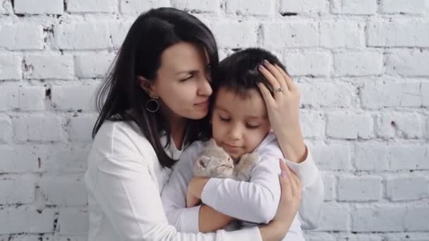 Vacker ung brunett mor i vita casual kläder kramar baby pojke på en vit bakgrund i vardagsrummet. En pojke på 5-6 år kramar en katt. Hem kramar, kärleksfull familj — Stockvideo