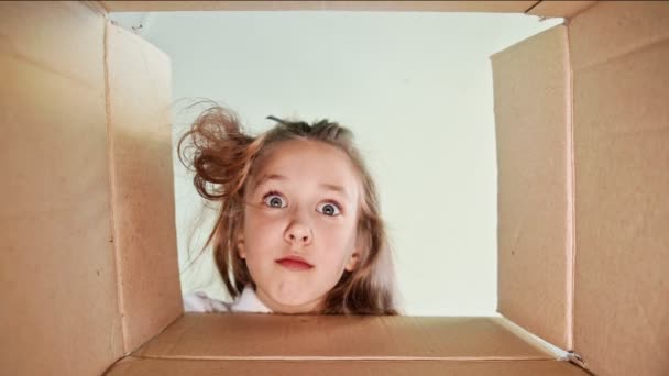 Seriöst barn, förtjust över en överraskning, öppnar en låda med gåvor. Porträtt av en vacker liten flicka, ett barn öppnar en låda med en överraskning ser in i en pappkartong för en gåva. - Inomhus. Långsamt — Stockvideo