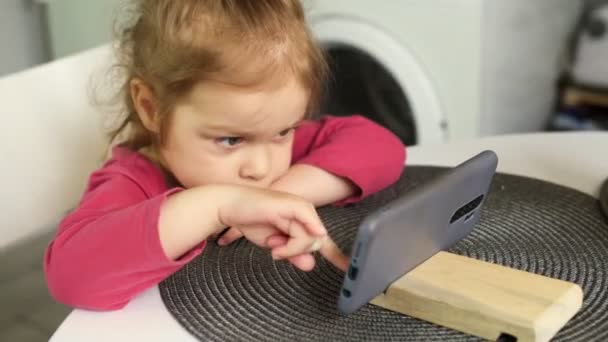 Small girl 2-3 years with a smartphone in her hands. Child play with phone.Girl at table play on smartphone. Child play games on phone. Happy little girl is play at home. Child sits at table and holds — 图库视频影像