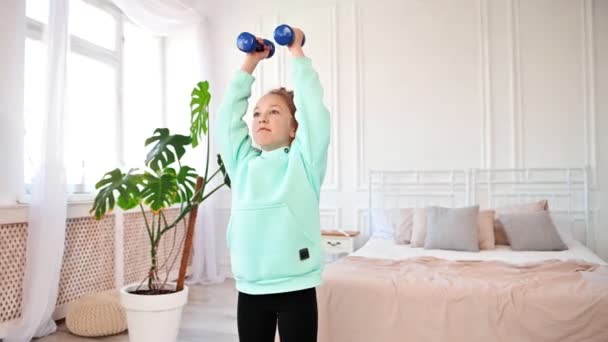 Concentrated little girl doing sports exercises with dumbbells in her hands. Teenagers and sports at home. — Video Stock