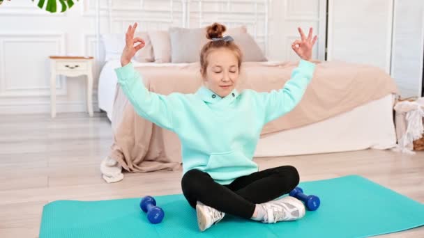 Aantrekkelijk klein meisje dat thuis yoga doet op een mat. Het kind zit in de lotuspositie. Langzame beweging — Stockvideo