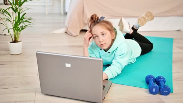 Das Mädchen denkt nach und sucht im Laptop nach Sportübungen. Baby Färse liegt neben Laptop im Wohnzimmer. Kinder und Sport. — Stockvideo