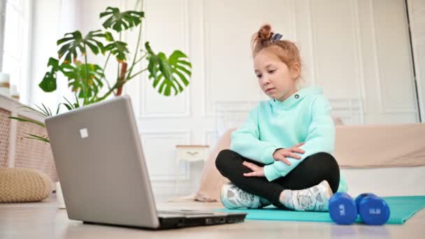 Meisje in de buurt van een laptop met verveling kijkend naar het scherm naast sportuitrusting. Leren op afstand lichamelijke opvoeding. Kinderen en technologie — Stockvideo