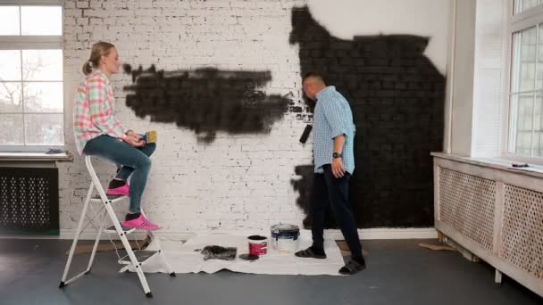 Schönes Paar schmückt seine neue Wohnung und albert herum. Mann und Frau bemalen die Wand mit Rollen, die in schwarze Farbe getaucht sind. — Stockvideo