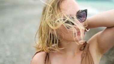 Güneş gözlüklü, sarı saçlı, deniz rüzgarı tarafından yakından çekilen çekici kız saçlarını uzatıyor. Yavaş çekim.