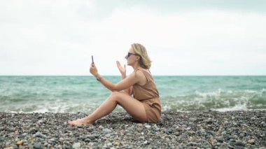 Kız videoda konuşuyor. Sahilde okyanusun arka planında oturuyor. Dalgalarla..