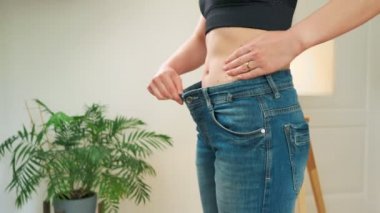 İnce belli, ince vücutlu genç fitness kadın, büyük pantolonlar deniyor ve evdeki mutfakta kameraya nasıl kilo verileceğini gösteriyor. Atletik kız diyetten sonra kilo verdi.