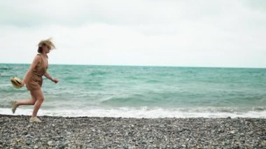 Slender attractive girl in light natural clothes runs on the beach along the frame on the background of the ocean with waves.