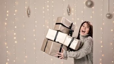 Süveter giymiş neşeli beyaz esmer, elinde bir yığın hediye, arka planda çelenkler var.