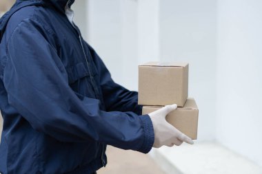 Paket teslimatı konsepti. Beyaz lastik eldivenli ve açık renkli koyu mavi ceketli postacı binanın önünde müşterisine küçük paketler vermek için bekliyor..