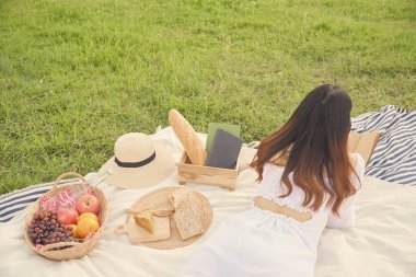 Piknik konsepti. Uzun kahverengi saçlı, rahat beyaz elbiseli kızın arkası. Piknik örtüsüne uzanıp kitap okumak..