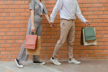 Alışveriş konsepti açık kahverengi pantolonlu ve beyaz ayakkabılı adam alışveriş çantasını solunda taşıyor ve gri pantolonlu ve siyah ayakkabılı kadının elini tutuyor..