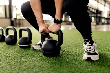 Egzersiz konsepti yeşil halının üzerine dizilmiş siyah kettlebell çanları gri ve siyah ayakkabılar giyen birinin yanında sallanıyor..