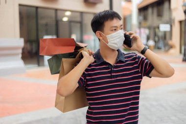 Alışveriş konsepti. Beyaz maske takan, arkadaşıyla telefonda konuşan ve sırtında alışveriş torbaları tutan adam.