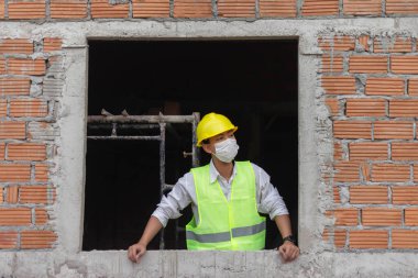 Mühendis konsepti. Canlı sarı bir şapka ve beyaz bir maske takan inşaat işçisi. Pencereden dışarı bakmaya çalışıyor..