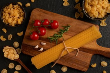 makarna konsepti çeşitli makarna çeşitleri küçük bir kaseye konuldu, bazıları tahta kaşığa kondu, bazıları kesim tahtasına konuldu, ve bazıları diğer malzemelerle birlikte koyu arka plana konuldu..