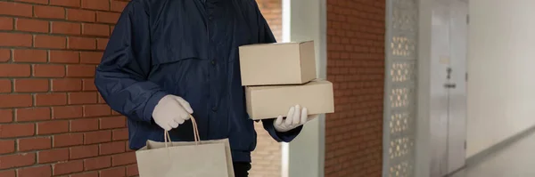 左手に袋一つと右手に箱二つを持ってこれらの荷物を彼の顧客にドロップする準備ができている薄い灰色のキャップを身に着けている背の高いポストキャリアを概念小包配達 — ストック写真