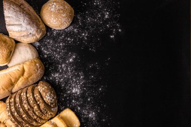Ekmek kavramı. Tohumlar, çörekler, kruvasanlar ve somun ekmekler ile birlikte birkaç dilim koyu kahverengi ekmek. Siyah arka planda toplanmış un tozu serpiştirilmiş..