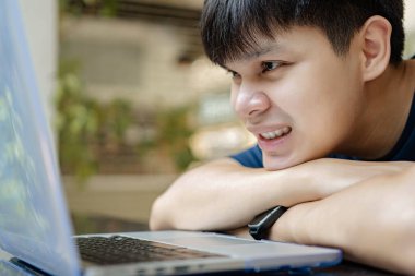 Çevrimiçi konsept üzerinde çalışan genç çocuk çenesini kollarına dayayıp dizüstü bilgisayara bakıyor, işine devam etmek için hiçbir fikri yok, bu da onu şu anda çalışmaktan bezdiriyor..