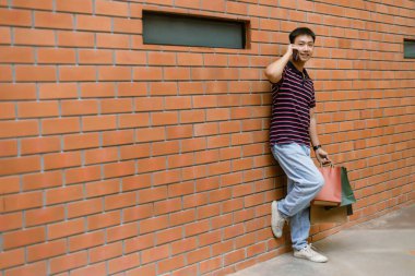 Alışveriş konsepti. Mutlu bir yüz ifadesi takınıp duvara yaslanmış ve yanında bir sürü alışveriş poşeti getirirken telefon etmeye çalışan adam..