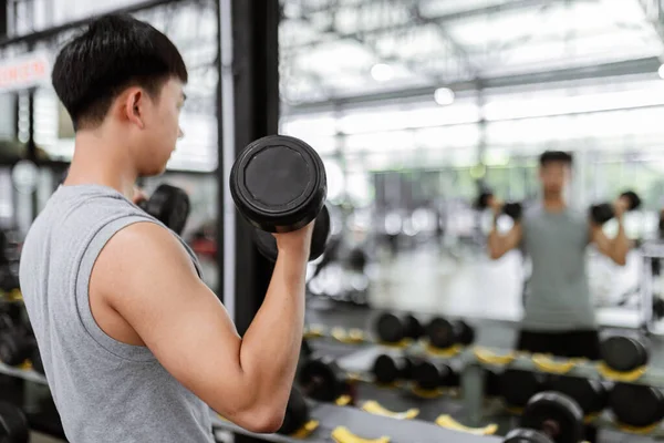 exercise concept The male sport member lift black dumbbells up with both hands, standing and doing concentration curl posture while looking at the mirror.