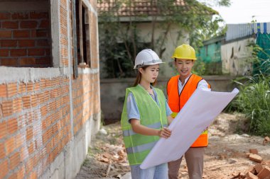 Mühendislik konsepti. Başlıklı ve maskeli iki bina yöneticisi binanın yanında duruyor ve binanın diyagramına bakıyor..