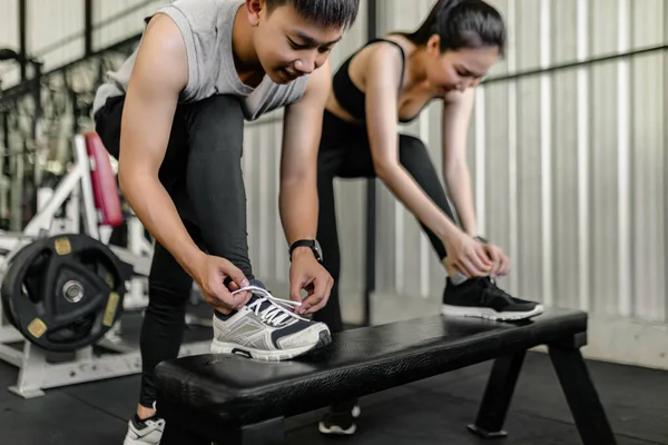 exercise concept The man and woman in the sport club resting one of their feet on the black bench and trying to tie up their shoelace.