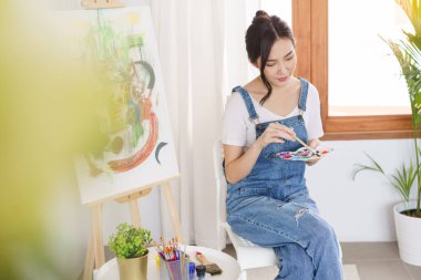 Sanat konsepti, genç Asyalı kadın boya fırçasını palete batırıp tuvale resim yapmak için kullanır..