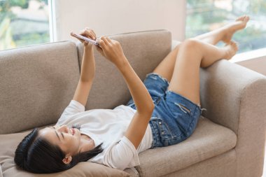 Rahat yaşam tarzı konsepti, kanepede uzanan genç bir kadın ve evde film izlemek için akıllı telefonu kullanmak..