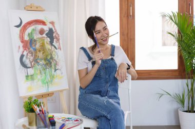 Sanat eserleri konsepti, genç Asyalı kadın boya fırçasını stüdyoda tuval üzerine resim eşliğinde ağzına koydu..