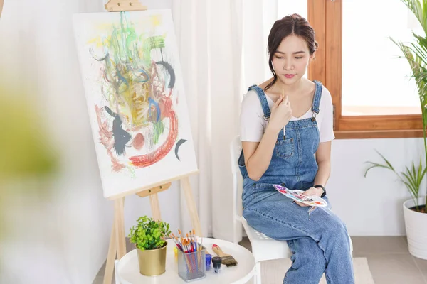 Sanat konsepti, genç Asyalı kadın boya fırçası tutuyor ve stüdyodaki sanat eserlerini düşünüyor..