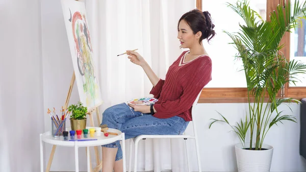Sanat konsepti, genç Asyalı kadın stüdyoda oturuyor ve elinde fırçayla sanat eserlerini işaret ediyor..
