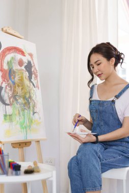 Sanat konsepti, genç Asyalı kadın boya fırçasını palete batırıp tuvale resim yapmak için kullanır..