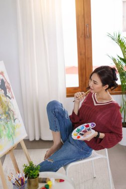 Sanat konsepti, genç Asyalı kadın boya fırçasını renk paletiyle tutuyor ve sanat eserlerine bakıyor..