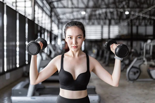 exercise concept The healthy woman in dark sport clothes tying her hair up and working out with weightlifting, carrying two black dumbbells up.