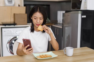 Evde rahatlama konsepti, kraker yiyen ve akıllı telefondan sosyal medyada sörf yapan genç bir kadın..