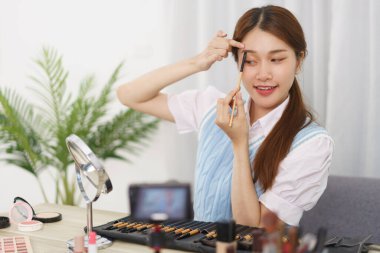 Beauty blogger concept, Young Asian woman is applying eyebrow to review cosmetic in live streaming.