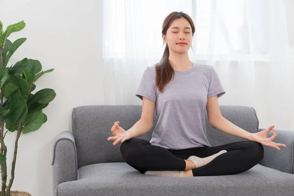 Yoga exercise concept, Young Asian woman doing yoga exercise in lotus pose on couch in living room.