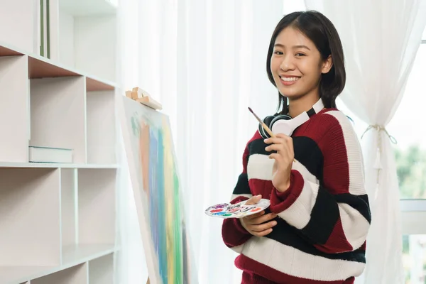 The art concept, Asian female artist holding brush color palette and smiling to happy with artwork.