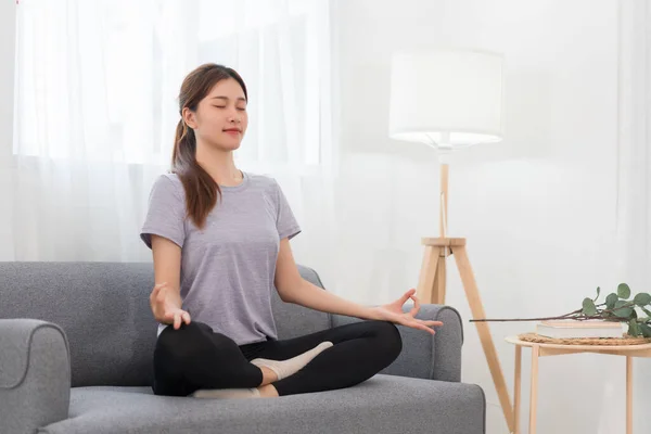 Yoga exercise concept, Young Asian woman doing yoga exercise in lotus pose on couch in living room.